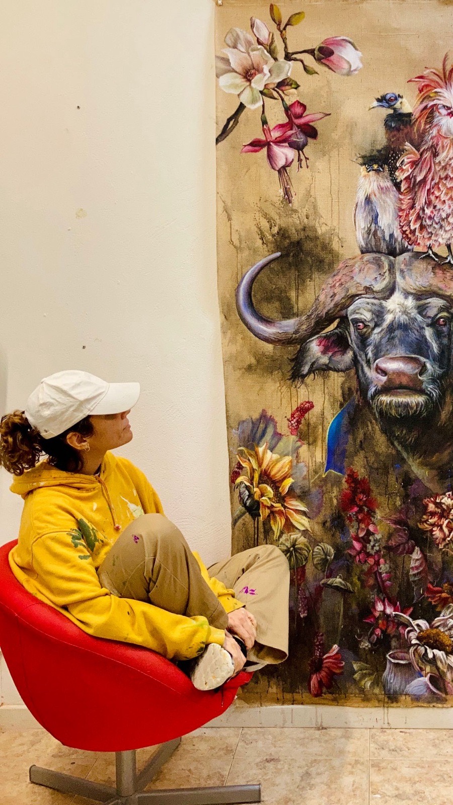 Lau Rodríguez sentada en un sillón rojo mirando una gran pintura con un búfalo, aves y flores